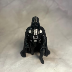 May include: A black figurine of a character wearing a helmet, a long cape, and a chest control panel with silver accents. The figure is posed in a kneeling position with one arm extended forward and the other resting near the knee.