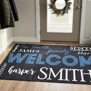 May include: A rectangular dark gray floor mat featuring the word WELCOME in large blue striped letters and the word SMITH in large white letters. The mat includes various names such as James, Jackson, Harper, and Sophia in different fonts.