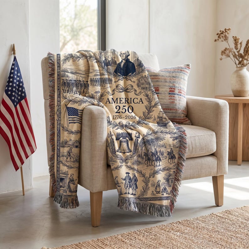 May include: A cream-colored throw blanket draped over a beige armchair. The blanket features a blue and white toile design with historical scenes, the American flag, and the text "AMERICA 250 1776-2026". The blanket has a fringed edge and is paired with a striped pillow.