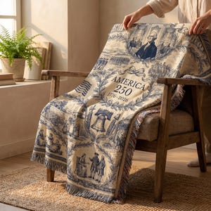 May include: A beige and blue throw blanket draped over a wooden chair. The blanket features historical illustrations and the text "AMERICA 250 1776-2026". The blanket has a fringed edge.