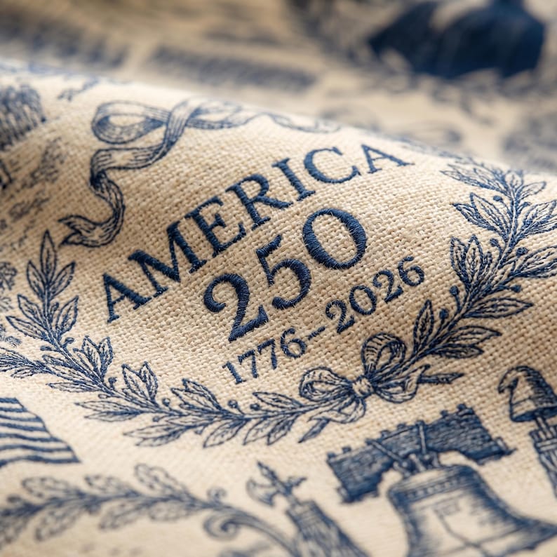 May include: Close-up of a beige fabric with a blue embroidered design. The text "AMERICA 250 1776-2026" is prominently displayed within a laurel wreath. Additional blue illustrations include the Liberty Bell and an American flag.