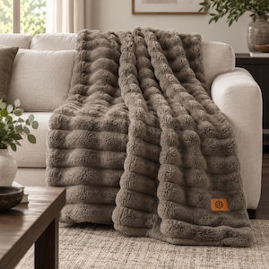 May include: A taupe faux fur throw blanket with a textured horizontal ribbed pattern. The blanket features a rectangular brown leather patch in the bottom corner with the monogram letters GZB embossed in a circular design.