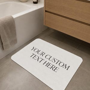 May include: A rectangular white bath mat with rounded corners featuring the black text YOUR CUSTOM TEXT HERE printed in the center. The mat has a flat surface and is positioned on a gray tiled floor in front of a bathroom vanity and bathtub.