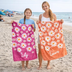 May include: Two beach towels with daisy patterns. One is pink with the name "Lily", the other is orange with the name "Laura". Both towels have white daisies with yellow centers. The towels are held up on a sandy beach.