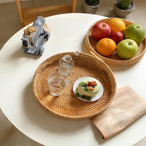 Könnte beinhalten: Zwei runde Tabletts aus Korbweide auf einem weißen Tisch. Eines enthält ein Glas Wasser, eine kleine Glasflasche und einen kleinen Kuchen auf einem Teller. Das andere enthält Äpfel, Orangen und eine grüne Frucht. Ein dekoratives Pferd und eine gerollte Zeitung sind ebenfalls vorhanden.