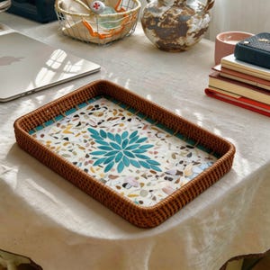 Puede incluir: Bandeja rectangular de ratán con diseño de mosaico. La bandeja presenta un borde tejido marrón, un ribete turquesa y un diseño floral central en tonos turquesa. El interior está relleno con un patrón estilo terrazo.