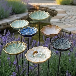 Könnte beinhalten: Eine Sammlung von sieben blumenförmigen Gartensteckern aus Keramik in verschiedenen Farben wie Hellgrün, Blau, Braun und Weiß. Jede schalenartige Blüte sitzt auf einem dünnen Metallstab. Eine weiße Blüte enthält eine Biene und klare Glasperlen.