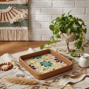 May include: A square woven tray with a mosaic floral design in shades of teal, purple, and cream. The tray is made of natural-colored wicker and sits on a patterned rug. A plant in a macrame holder is in the background.