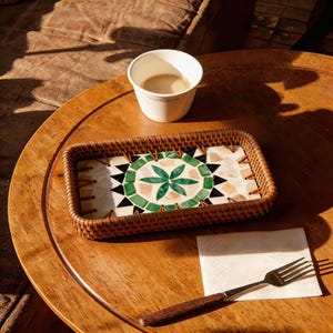 May include: A rectangular woven tray with a mosaic tile design featuring a green floral pattern, black triangles, and beige tiles. A white paper cup with coffee sits on a wooden table, along with a napkin and a fork.