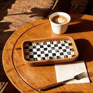 Puede incluir: Una bandeja rectangular tejida con un patrón de cuadros blancos y negros se encuentra sobre una mesa de madera. Una taza de café con arte latte, una servilleta y un tenedor también están sobre la mesa. La bandeja tiene un borde tejido y detalles dorados.