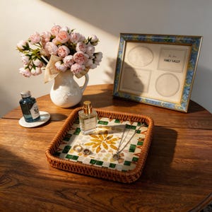Puede incluir: Una bandeja decorativa con un diseño de mosaico contiene joyas y perfume. Un ramo de flores rosas está en un jarrón blanco, y una foto enmarcada con el texto "FAMILY GAILLY" está sobre una mesa de madera.