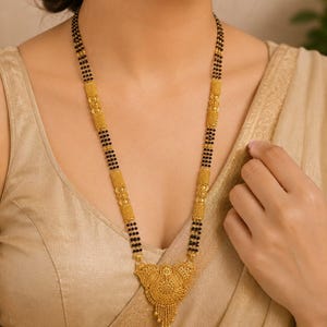 Op de afbeelding: Een lange gouden ketting met een reeks kleine zwarte kralen afgewisseld met cilindrische, getextureerde gouden tussenstukjes. De ketting eindigt in een grote, driehoekige, gedetailleerde gouden hanger met een filigraanontwerp en kleine bungelende kraaltjes.
