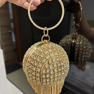 Puede incluir: Un bolso de noche redondo dorado, cubierto de brillantes pedrería. El bolso presenta un asa grande y circular y una borla de pequeñas cuentas doradas. La superficie del bolso está densamente cubierta de pedrería.