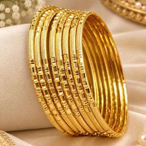 May include: A set of seven gold-toned bangles featuring a polished finish with alternating textured segments. Each circular bracelet displays a repeating pattern of smooth, reflective sections and finely etched, matte-finished rectangular details.