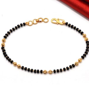 May include: Gold-toned bracelet featuring alternating black beads and gold-colored, textured beads. The bracelet has a clasp closure and a series of small gold rings at the other end. The jewelry is displayed on a white surface.