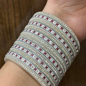 May include: A stack of white bangles with a repeating pattern of red crosses and small green dots. The bangles are also adorned with rows of small, sparkling rhinestones. The design is set against a cream-colored background.