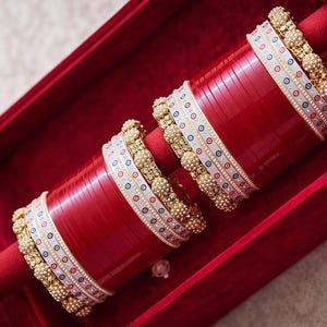 Può includere: Un set di braccialetti rossi con dettagli bianchi e dorati, presentato in una scatola di velluto rosso. I braccialetti presentano dettagli intricati con piccole perle e motivi circolari colorati. Un gioiello tradizionale.