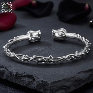 Könnte beinhalten: Silberfarbenes Armreif-Armband mit floralem und dornigem Design. Das Armband hat rosenförmige Enden und ein detailliertes Muster aus Ranken, Blättern und Dornen. Der Schmuck wird auf einer dunklen, strukturierten Oberfläche präsentiert.