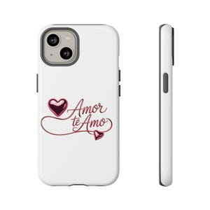 Peut inclure: Coque de téléphone blanche avec un motif en forme de cœur et les mots "Amor te Amo" en écriture cursive. Le cœur et le texte sont de couleur rouge métallique. La coque a un objectif de caméra et des boutons noirs.
