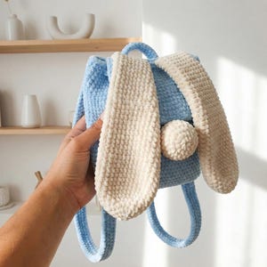 Puede incluir: Mochila de ganchillo azul claro con forma de conejito. Tiene largas orejas color crema y una cola de pompón color crema. La mochila tiene un asa y dos correas.
