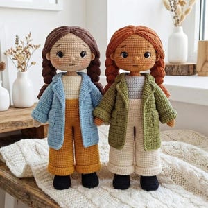 Puede incluir: Dos muñecas de ganchillo hechas a mano. Una muñeca tiene el pelo castaño en trenzas, con una chaqueta azul claro, pantalones mostaza y zapatos negros. La otra muñeca tiene el pelo rojo en trenzas, con una chaqueta verde, pantalones crema y zapatos negros.