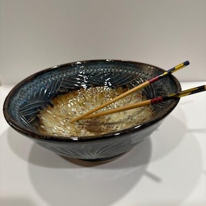 Könnte beinhalten: Eine handgefertigte Keramikschale mit dunkelblauem Rand und strukturiertem, hellbraunem Innenraum. Zwei Essstäbchen aus Holz mit farbenfrohen Akzenten liegen in der Schale. Die Schale hat eine einzigartige, organische Form, geeignet für Suppen oder Nudeln.
