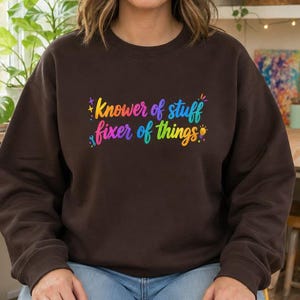 May include: A dark brown crewneck sweatshirt featuring the colorful text "Knower of stuff fixer of things" printed on the chest. The design includes small graphic icons of a wrench, a lightbulb, and stars in a vibrant rainbow gradient.