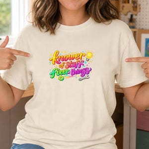 Può includere: Una t-shirt a maniche corte color crema con una stampa grafica colorata sul petto. Il testo "Knower of Stuff Fixer of things" è scritto in un vivace carattere corsivo multicolore, accompagnato da una lampadina gialla, un piccolo ingranaggio e una chiave inglese.