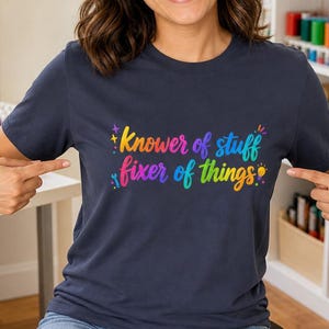 May include: A dark navy blue short-sleeve t-shirt featuring the multi-colored text "Knower of stuff fixer of things" printed on the chest. The design includes small graphic icons of a wrench, a lightbulb, and stars in vibrant rainbow colors.