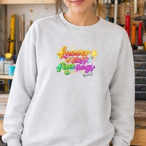 May include: A light grey crewneck sweatshirt featuring a colourful graphic on the chest. The text reads "Knower of Stuff" in yellow and pink, and "Fixer of things" in green and purple, accompanied by a yellow lightbulb, a green gear, and a grey wrench.