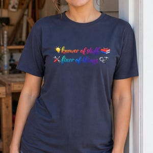 Könnte beinhalten: Ein dunkelblaues T-Shirt mit kurzen Ärmeln und einem mehrfarbigen Grafikdruck. Der Text lautet "knower of stuff" mit einer Glühbirne und Büchern sowie "fixer of things" mit gekreuzten Schraubendrehern, einem Zahnrad und einem Hammer.