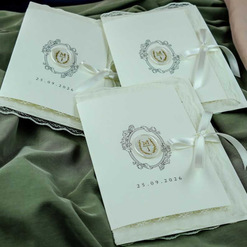 K&ouml;nnte beinhalten: Drei cremewei&szlig;e, rechteckige Hochzeitseinladungen mit einem schwarzen, verzierten ovalen Rahmen, einem zentralen wei&szlig;en Wachssiegel mit goldenen Initialen M und I, einer wei&szlig;en Satinschleife und dem Datum 25.09.2026, verziert mit wei&szlig;er Spitze.