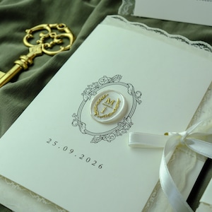 K&ouml;nnte beinhalten: Eine cremefarbene Einladungskarte mit einer schwarzen, verzierten Rahmenillustration, einem wei&szlig;en Wachssiegel mit einem goldenen Buchstaben M und dem in Schwarz gedruckten Datum 25.09.2026. Die Karte ist mit einem wei&szlig;en Satinband und einem goldenen Schl&uuml;ssel verziert.