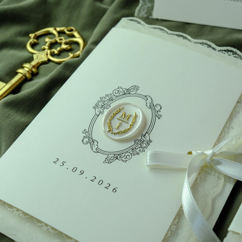 K&ouml;nnte beinhalten: Eine cremefarbene Einladungskarte mit einer schwarzen, verzierten Rahmenillustration, einem wei&szlig;en Wachssiegel mit einem goldenen Buchstaben M und dem in Schwarz gedruckten Datum 25.09.2026. Die Karte ist mit einem wei&szlig;en Satinband und einem goldenen Schl&uuml;ssel verziert.