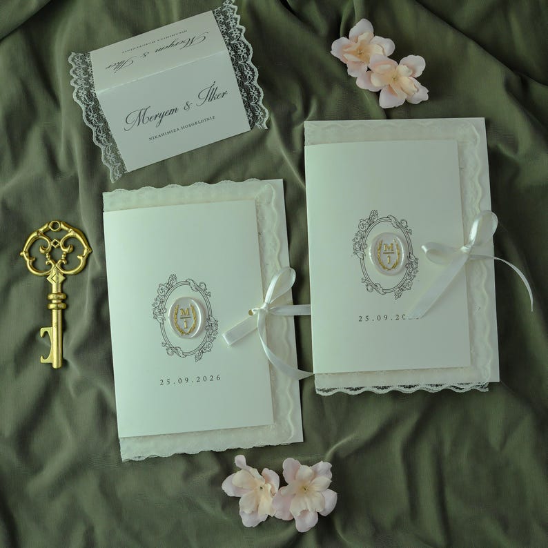 K&ouml;nnte beinhalten: Zwei cremefarbene Hochzeitseinladungskarten mit wei&szlig;er Spitzenbord&uuml;re, jeweils mit einem zentralen Wachssiegel mit den Initialen MI, einem wei&szlig;en Satinband und aufgedruckten Daten. Ein goldfarbener Skelettschl&uuml;ssel und zwei blassrosa Kunstblumen.