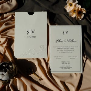 Peut inclure: Ensemble de faire-part de mariage ivoire avec des détails floraux en relief. L'invitation comprend la date 13/06/2026 et les noms Lilan & Volkan. L'ensemble est présenté sur un fond en tissu drapé avec une clé et des fleurs.