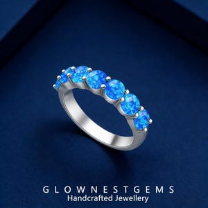 Puede incluir: Un anillo plateado con seis gemas redondas de color azul brillante. El anillo se encuentra sobre un fondo azul oscuro. El texto "GLOWNESGEMS Handcrafted Jewellery" está en la parte inferior de la imagen.