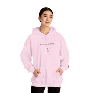 Puede incluir: Sudadera con capucha rosa claro con cordón y bolsillo delantero. El texto "don't trip the crip" y un dibujo de un hombre de palo están impresos en la parte delantera. La sudadera está hecha de tela suave.