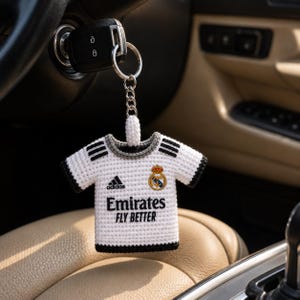 Può includere: Un portachiavi all'uncinetto bianco e nero a forma di maglia da calcio. La maglia presenta il logo Adidas, il logo Emirates Fly Better e uno stemma della squadra. Il portachiavi è attaccato a una chiave dell'auto.