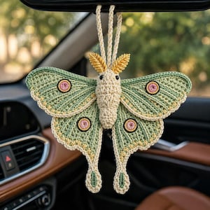 Puede incluir: Una polilla hecha a mano con ganchillo en tonos verdes, crema y amarillo, colgada del espejo retrovisor de un coche. La polilla presenta patrones de alas detallados, detalles circulares rosas y negros y un cuerpo color crema. Las alas miden unos 20 cm de ancho.