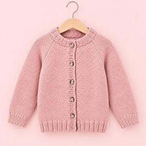 Può includere: Un cardigan in maglia a maniche lunghe color rosa polvere con girocollo, polsini e orlo a coste. Il capo si chiude sul davanti con cinque bottoni rotondi marroni maculati ed è appeso a una gruccia di legno.