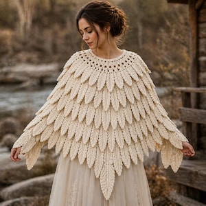 Puede incluir: Capa de ganchillo color crema con un diseño en forma de pluma. La capa presenta un escote redondo y elementos en forma de hoja que caen en cascada sobre los hombros y los brazos. La prenda se lleva sobre un vestido color crema.