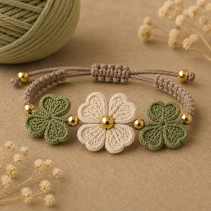 May include: A macrame bracelet featuring a central cream-colored flower motif flanked by two green clover-shaped accents. The design includes small gold-toned metallic beads between the floral elements and a tan adjustable cord band.