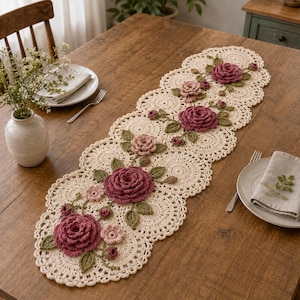 May include: An elongated cream-coloured crochet table runner featuring a series of circular motifs with scalloped edges. The runner is decorated with three-dimensional dark pink and light pink crocheted roses and green crocheted leaves.