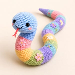 May include: A crocheted snake toy in rainbow colours with a smiling blue head, pink cheeks, and a red tongue. The body is striped and decorated with embroidered flowers and a crescent moon.