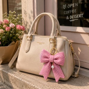 May include: Cream-colored handbag with top handles and a detachable shoulder strap. The bag features gold-tone hardware and a pink crocheted bow keychain with a pearl accent. The text "DREAM & LOVE" is visible on the bag.