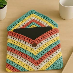 May include: A rectangular crochet envelope sleeve featuring a chevron pattern in teal, yellow, cream, and terracotta yarn. The sleeve has a pointed flap secured by a single brown button, containing a black tablet device inside the main compartment.