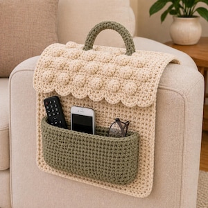May include: A cream-colored, crocheted sofa arm organizer with a green pocket and handle. The organizer has a scalloped edge and a textured, dotted pattern. A remote, phone, and glasses are in the pocket.