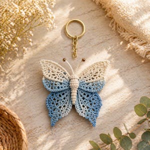 May include: A handmade crochet butterfly keychain in shades of blue and cream. The butterfly has a detailed openwork design and is attached to a gold-colored keyring. The wings transition from cream to blue. Decorative item.