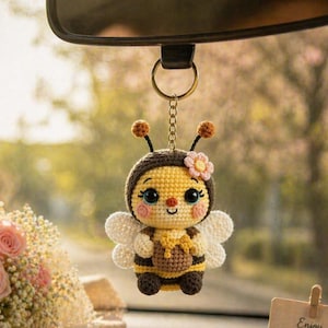 May include: A crocheted bee keychain with a yellow and brown body, white wings, and a flower on its head. The bee has large blue eyes and is hanging from a gold-colored chain and ring. The background is blurred, suggesting an outdoor setting.